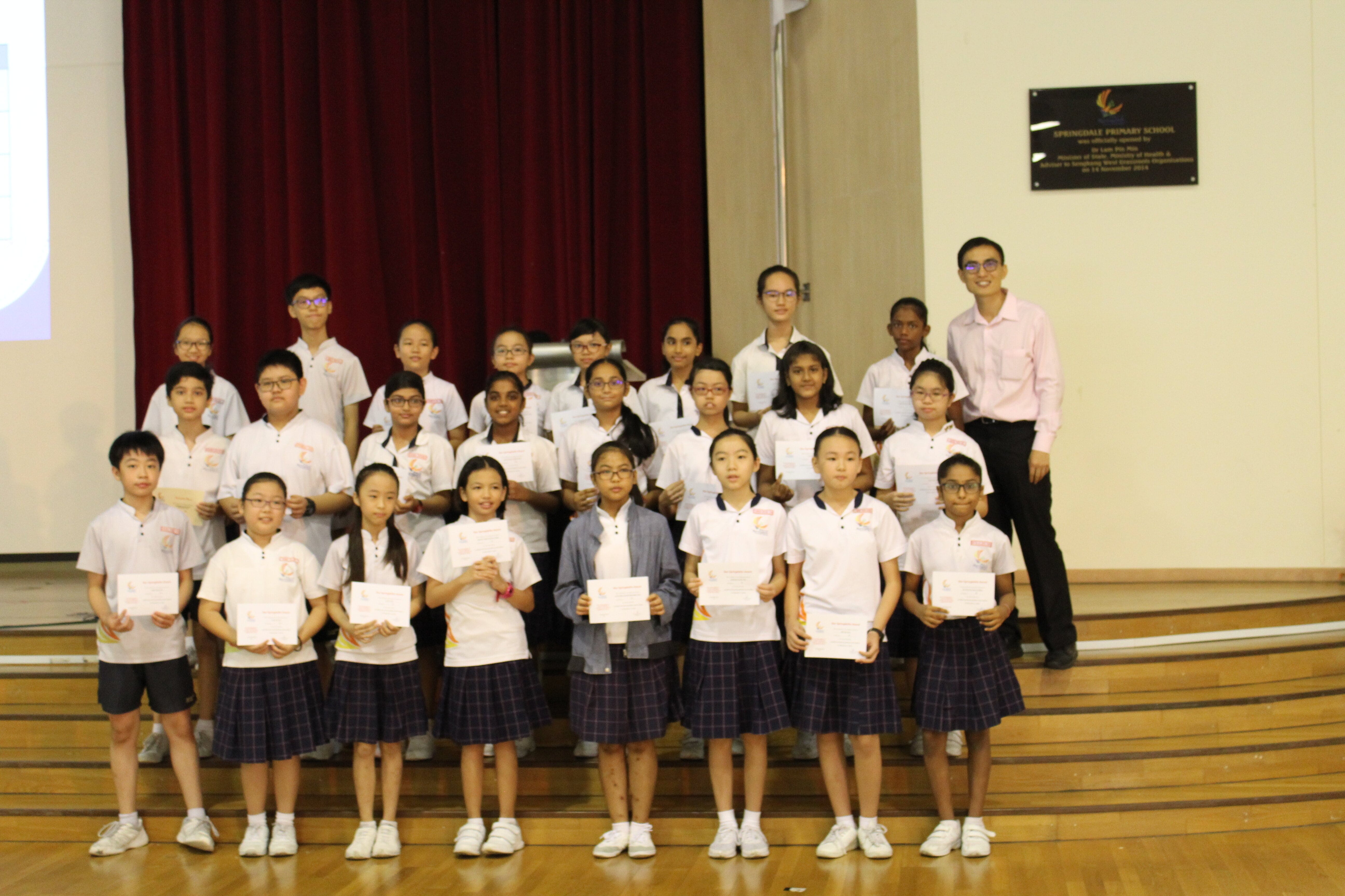 A group of primary school students in uniforms holding certificates, posing with a teacher on a school stage.